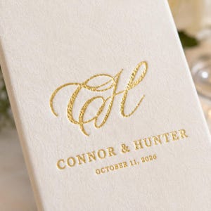 K&ouml;nnte beinhalten: Eine cremefarbene, strukturierte Karte mit einem goldfoliengepr&auml;gten Monogramm des Buchstabens H in Schreibschrift. Unter dem Monogramm steht der Text CONNOR & HUNTER sowie das Datum OCTOBER 11, 2026, ebenfalls in goldener Schrift.