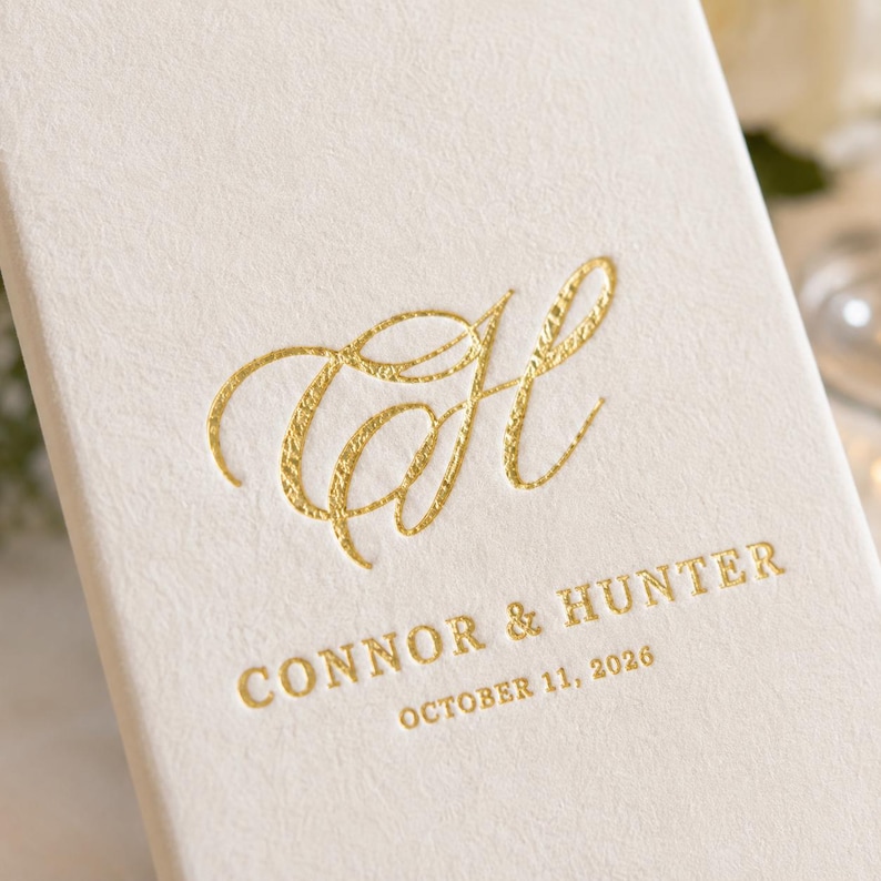 K&ouml;nnte beinhalten: Eine cremefarbene, strukturierte Karte mit einem goldfoliengepr&auml;gten Monogramm des Buchstabens H in Schreibschrift. Unter dem Monogramm steht der Text CONNOR & HUNTER sowie das Datum OCTOBER 11, 2026, ebenfalls in goldener Schrift.