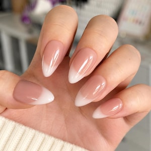 May include: A hand showing long, almond-shaped nails with a French manicure. The nails have a sheer, pink-nude base with white tips. The manicure is glossy and well-maintained, presenting a clean and elegant style, suitable for various occasions.