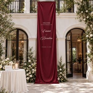 Può includere: Un lungo striscione verticale in tessuto bordeaux scuro appeso a una parete bianca. Lo striscione presenta un testo bianco che recita: WELCOME TO THE WEDDING OF Emma & Brendan November 13, 2026. Il tessuto ha una finitura liscia e opaca con pieghe.