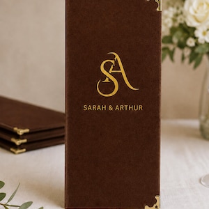 K&ouml;nnte beinhalten: Ein rechteckiger Men&uuml;kartenhalter aus braunem Samt mit einem goldenen Folienmonogramm der Buchstaben SA und dem Schriftzug SARAH & ARTHUR. Der Einband ist an den oberen und unteren rechten Ecken mit polierten goldfarbenen Metallecken versehen.