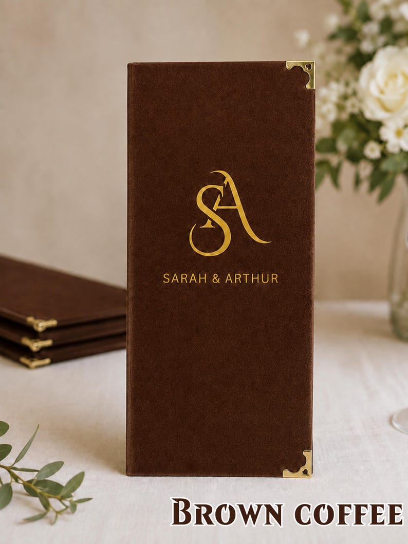 K&ouml;nnte beinhalten: Ein rechteckiger Men&uuml;kartenhalter aus braunem Samt mit einem goldenen Folienmonogramm der Buchstaben SA und dem Schriftzug SARAH & ARTHUR. Der Einband ist an den oberen und unteren rechten Ecken mit polierten goldfarbenen Metallecken versehen.