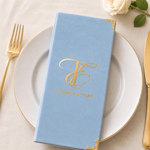 May include: A light blue rectangular menu cover with a soft texture, featuring gold foil corner accents and a gold foil monogram of the letters E and T, with the text ELIZABETH & TYLER printed in gold below the monogram on the front cover.