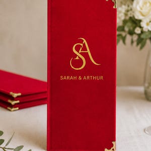 K&ouml;nnte beinhalten: Ein rechteckiger Men&uuml;einband aus rotem Samt mit goldfarbenen Metallecken. Die Vorderseite ist mit einem goldfarbenen Monogramm der Buchstaben SA und dem darunter zentrierten Text SARAH & ARTHUR bedruckt.