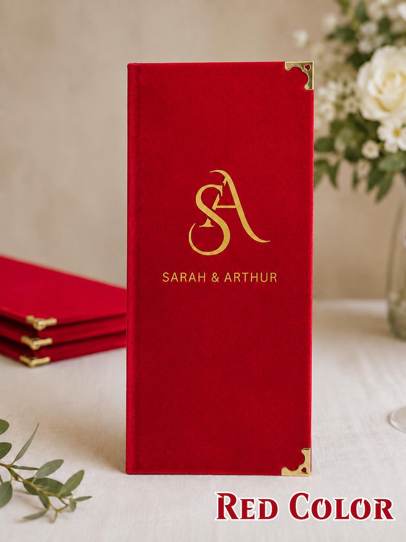 K&ouml;nnte beinhalten: Ein rechteckiger Men&uuml;einband aus rotem Samt mit goldfarbenen Metallecken. Die Vorderseite ist mit einem goldfarbenen Monogramm der Buchstaben SA und dem darunter zentrierten Text SARAH & ARTHUR bedruckt.