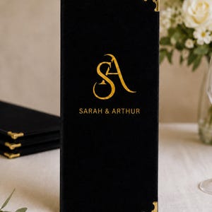 K&ouml;nnte beinhalten: Eine rechteckige schwarze Men&uuml;karte mit goldenen Metalleckensch&uuml;tzern. Auf der Vorderseite befindet sich ein goldenes Monogramm der Buchstaben SA &uuml;ber dem gedruckten Text SARAH & ARTHUR in einer serifenlosen Schrift auf der schwarzen Fl&auml;che.