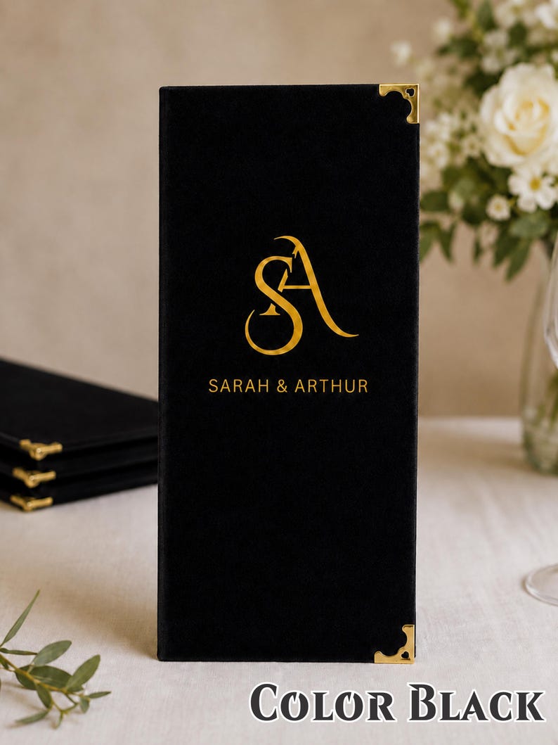 K&ouml;nnte beinhalten: Eine rechteckige schwarze Men&uuml;karte mit goldenen Metalleckensch&uuml;tzern. Auf der Vorderseite befindet sich ein goldenes Monogramm der Buchstaben SA &uuml;ber dem gedruckten Text SARAH & ARTHUR in einer serifenlosen Schrift auf der schwarzen Fl&auml;che.