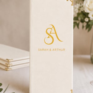K&ouml;nnte beinhalten: Ein vertikales, elfenbeinwei&szlig;es, rechteckiges Men&uuml; oder G&auml;stebuch mit goldenen Metalleckensch&uuml;tzern. Die Vorderseite zeigt ein goldenes Monogramm der Buchstaben S und A sowie den Text SARAH & ARTHUR in goldener serifenloser Schrift darunter.