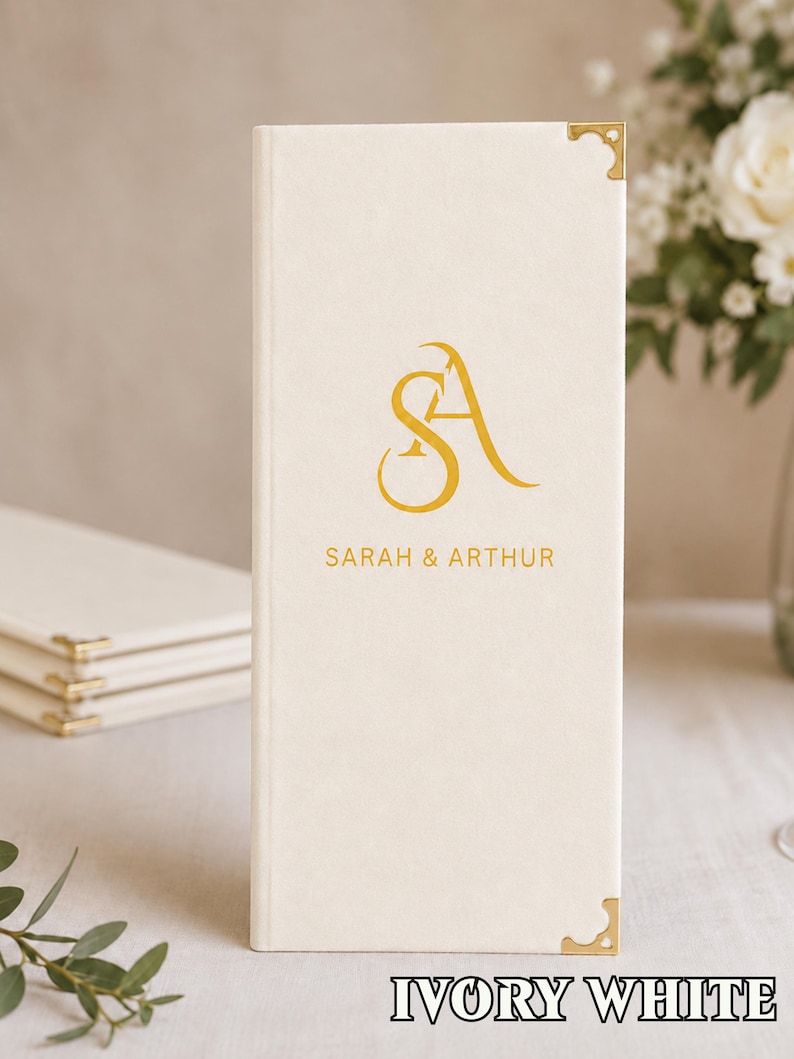 K&ouml;nnte beinhalten: Ein vertikales, elfenbeinwei&szlig;es, rechteckiges Men&uuml; oder G&auml;stebuch mit goldenen Metalleckensch&uuml;tzern. Die Vorderseite zeigt ein goldenes Monogramm der Buchstaben S und A sowie den Text SARAH & ARTHUR in goldener serifenloser Schrift darunter.