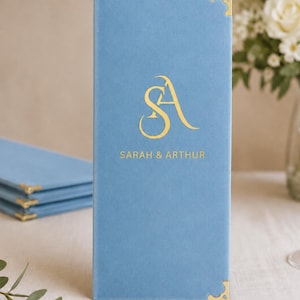 K&ouml;nnte beinhalten: Ein hellblaues, rechteckiges Men&uuml;cover mit weicher Textur und goldenen Metallecken. Auf der Vorderseite befindet sich ein goldenes Monogramm der Buchstaben SA &uuml;ber dem aufgedruckten Text SARAH & ARTHUR in einer serifenlosen Schrift.