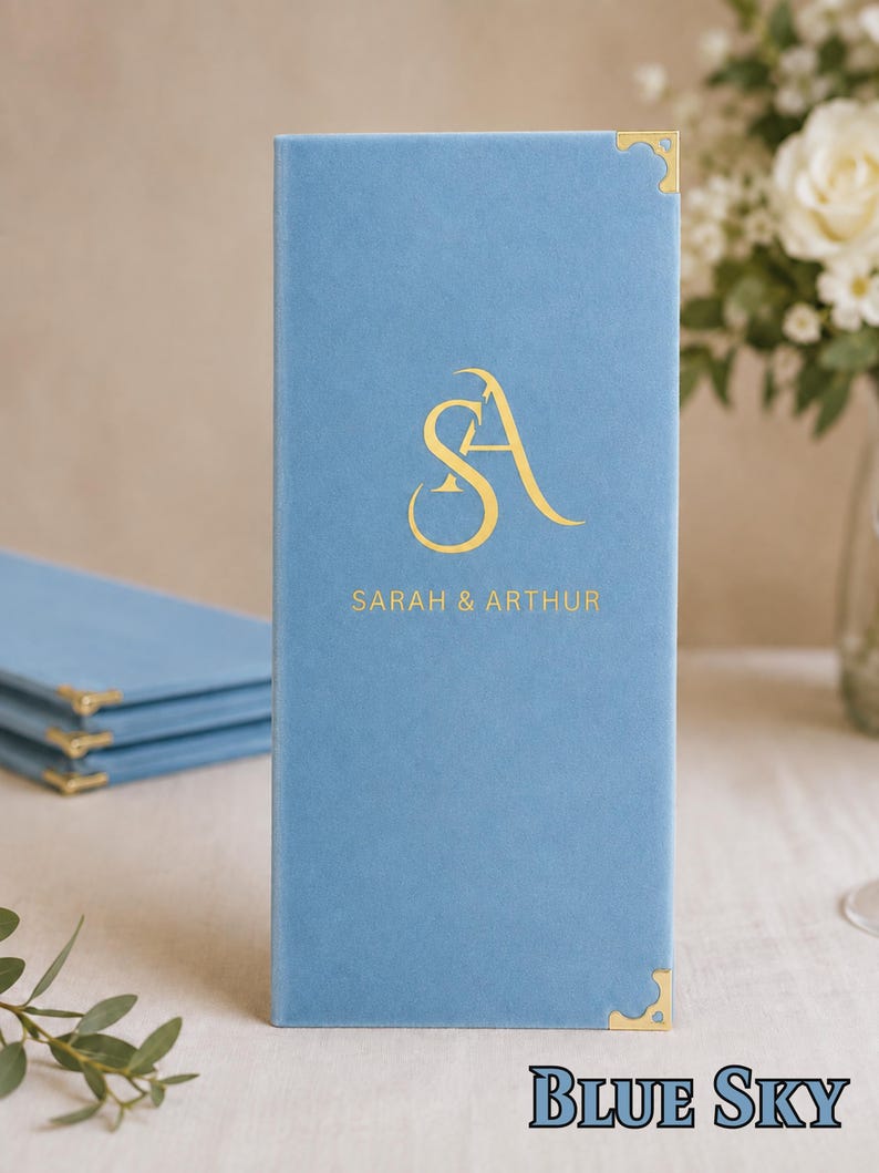 K&ouml;nnte beinhalten: Ein hellblaues, rechteckiges Men&uuml;cover mit weicher Textur und goldenen Metallecken. Auf der Vorderseite befindet sich ein goldenes Monogramm der Buchstaben SA &uuml;ber dem aufgedruckten Text SARAH & ARTHUR in einer serifenlosen Schrift.