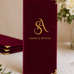 May include: A vertical rectangular menu cover in deep dark red velvet featuring gold metal corner protectors. The front displays a gold foil monogram of the letters SA above the printed names SARAH & ARTHUR in a clean, capitalized sans-serif font.