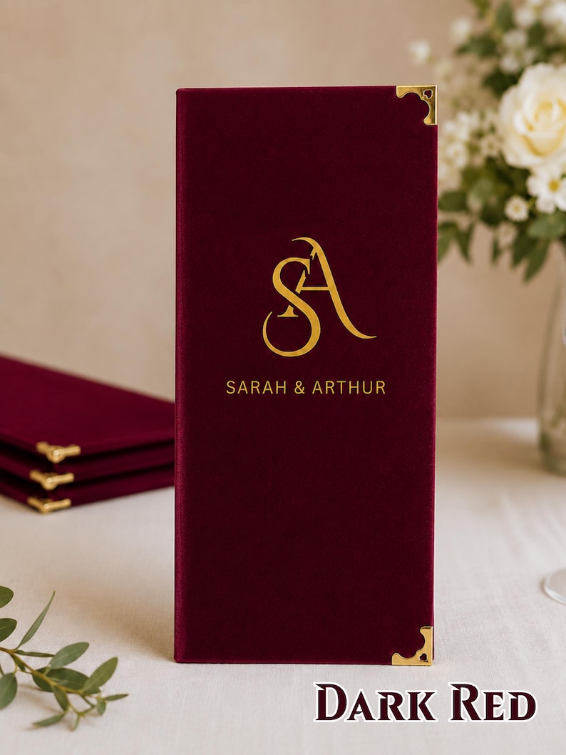 K&ouml;nnte beinhalten: Ein rechteckiger Men&uuml;einband aus dunkelrotem Samt mit goldenen Metalleckensch&uuml;tzern. Die Vorderseite zeigt ein goldenes Monogramm der Buchstaben SA &uuml;ber dem gedruckten Text SARAH & ARTHUR in einer zentrierten, serifenlosen Schriftart.