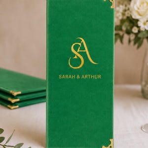 K&ouml;nnte beinhalten: Eine hohe, rechteckige Men&uuml;karte aus gr&uuml;nem Samt mit goldfarbenen Metallecken. Die Vorderseite ist mit einem goldfarbenen Monogramm der Buchstaben SA und dem darunter zentrierten Text SARAH & ARTHUR bedruckt.