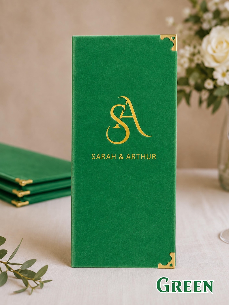 K&ouml;nnte beinhalten: Eine hohe, rechteckige Men&uuml;karte aus gr&uuml;nem Samt mit goldfarbenen Metallecken. Die Vorderseite ist mit einem goldfarbenen Monogramm der Buchstaben SA und dem darunter zentrierten Text SARAH & ARTHUR bedruckt.