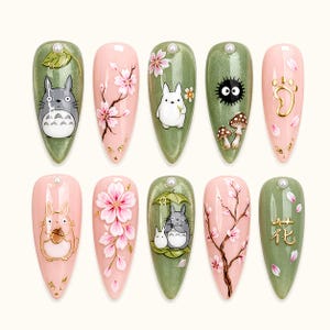 May include: A set of ten almond-shaped artificial nails with various designs. The designs include cartoon characters, cherry blossoms, and gold accents on a pink and green background. Each nail has a pearl at the base.