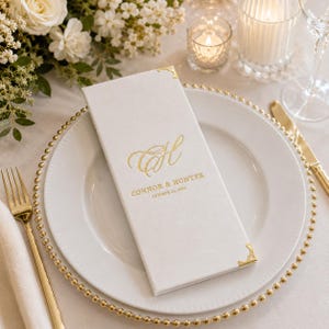 K&ouml;nnte beinhalten: Eine rechteckige wei&szlig;e Men&uuml;karte mit Goldfolienakzenten, die das Monogramm CH, die Namen Connor & Hunter sowie das Datum 13. Oktober 2026 zeigt. Die Karte hat dekorative Metallecken in Gold und liegt auf einem wei&szlig;en Speiseteller.