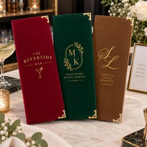 Könnte beinhalten: Drei hohe, rechteckige Menükartenhüllen aus samtigem Stoff in Dunkelrot, Waldgrün und Braun. Jede hat goldfarbene Metallecken und goldene Schriftzüge. Die rote Hülle zeigt THE RIVERSIDE BAR, die grüne M+K GOOD DRINKS BETTER COMPANY und die braune SL COCKTAILS PEOPLE MEMORIES.