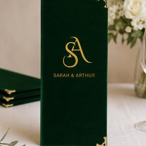 K&ouml;nnte beinhalten: Ein rechteckiger Men&uuml;einband aus dunkelgr&uuml;nem Samt mit einem goldenen Folienmonogramm der Buchstaben SA und dem Text SARAH & ARTHUR. Der Einband verf&uuml;gt &uuml;ber dekorative Metalleckensch&uuml;tzer in Gold an der oberen und unteren rechten Kante.