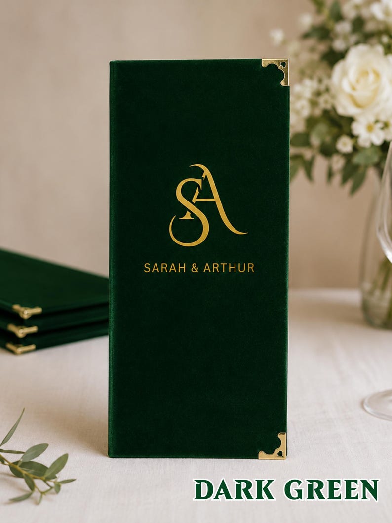 K&ouml;nnte beinhalten: Ein rechteckiger Men&uuml;einband aus dunkelgr&uuml;nem Samt mit einem goldenen Folienmonogramm der Buchstaben SA und dem Text SARAH & ARTHUR. Der Einband verf&uuml;gt &uuml;ber dekorative Metalleckensch&uuml;tzer in Gold an der oberen und unteren rechten Kante.