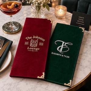 May include: Two rectangular velvet menu covers, one deep red and one dark green, each featuring gold-toned metal corner protectors. The red cover displays "The Johnson Lounge EST. 2026" with a cocktail graphic, while the green cover shows "ELIZABETH & TYLER" with a monogram.