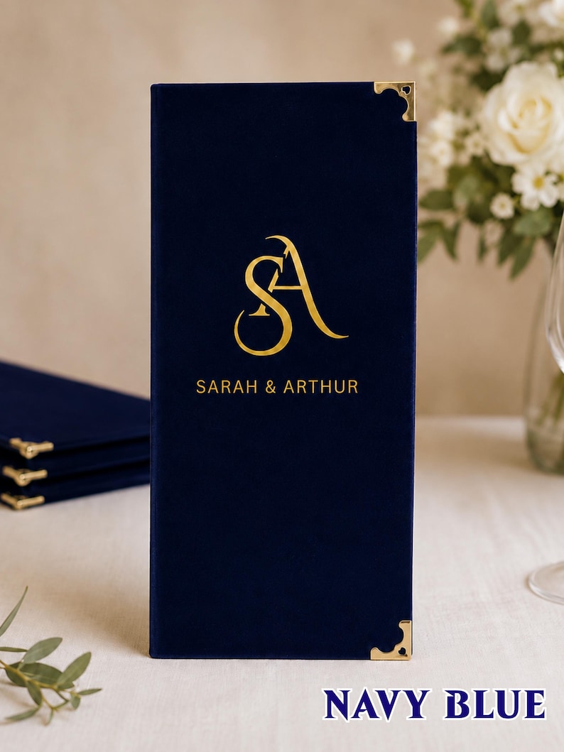 K&ouml;nnte beinhalten: Ein rechteckiger Men&uuml;einband aus marineblauem Samt mit goldenen Metalleckensch&uuml;tzern. Die Vorderseite ist mit einem goldenen Folienmonogramm der Buchstaben SA und dem darunter zentrierten Text SARAH & ARTHUR in einer serifenlosen Schrift versehen.