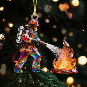 Puede incluir: Adorno de bombero facetado y colorido. La figura sostiene una manguera, rociando agua sobre una llama facetada. El nombre "Michael" está impreso en la parte posterior del adorno. El adorno cuelga de una cuerda dorada.