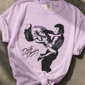 May include: A light purple short-sleeve t-shirt featuring a black graphic print of a couple performing a dance lift. The text Dirty Dancing is printed in black script below the figures. The shirt has a crew neckline and a Comfort Colors brand label.