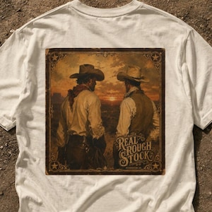 Peut inclure: Un t-shirt blanc cassé avec une impression graphique rectangulaire représentant deux cow-boys portant des chapeaux dans un paysage désertique au coucher du soleil. L'imprimé présente des bordures ornées d'étoiles et le texte "REAL ROUGH STOCK".