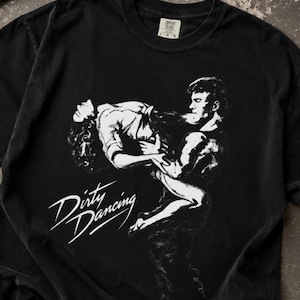 May include: A black crew neck t-shirt featuring a white graphic print of two people dancing, with the text Dirty Dancing written in a stylized script font. The shirt has a tag at the neckline indicating size large and the brand name Comfort Colors.