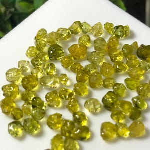 Könnte beinhalten: Eine Ansammlung kleiner, roher, durchscheinender gelb-grüner Edelsteine mit unregelmäßigen, facettierten Formen. Die Steine variieren in Größe und Farbintensität, von blassem Limettengrün bis zu dunklem Oliv, und liegen auf einer weißen Oberfläche.