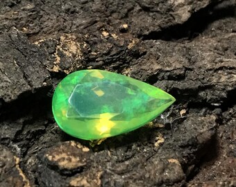 May include: A pear-shaped faceted gemstone displaying a vibrant green color with internal flashes of yellow and orange light. The stone features a smooth, polished surface with multiple geometric facets that reflect light across its translucent body.