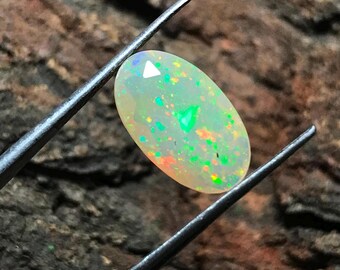 May include: An oval-shaped opal gemstone featuring a translucent, milky white base with vibrant flashes of green, orange, and red play-of-color. The gemstone is held by a pair of thin, silver-toned metal tweezers, showcasing its faceted surface.