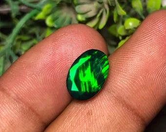 May include: An oval-shaped, faceted gemstone displaying a deep black base color with vibrant, luminous neon green flashes across its surface. The stone features a polished finish and a distinct play-of-color effect that shifts with light reflection.