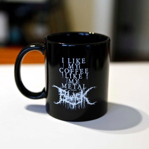 May include: Black coffee mug with a curved handle. The mug features white text that reads "I LIKE MY COFFEE LIKE MY METAL" with a stylized band logo below. The mug is made of ceramic.
