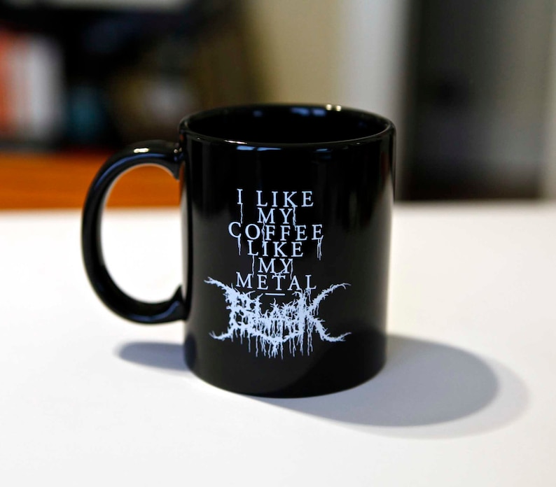 May include: Black coffee mug with a curved handle. The mug features white text that reads "I LIKE MY COFFEE LIKE MY METAL" with a stylized band logo below. The mug is made of ceramic.