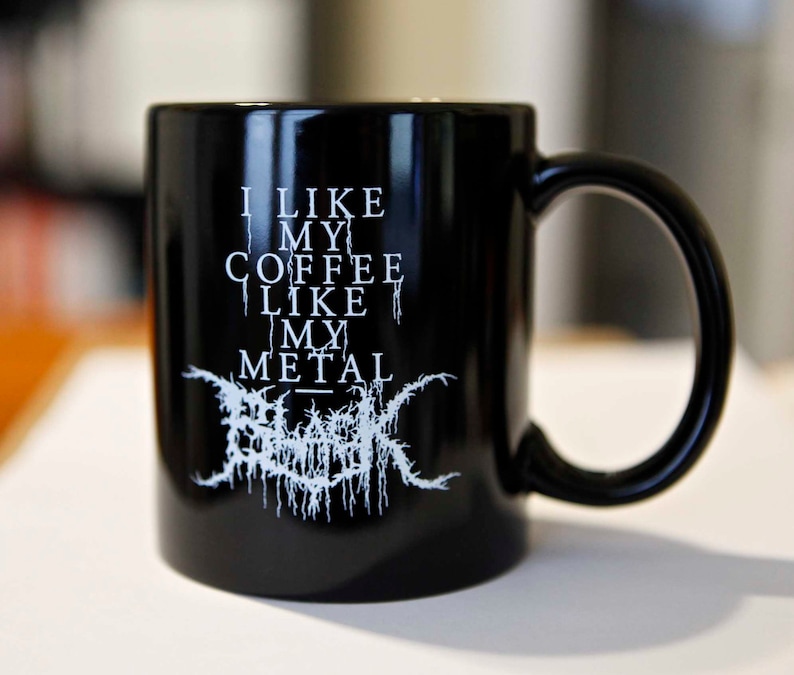 May include: Black ceramic coffee mug with a large handle. The mug features white text that reads "I LIKE MY COFFEE LIKE MY METAL" with a stylized logo below. The mug is designed for hot beverages.