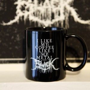 May include: Black ceramic mug with the text "I LIKE MY COFFEE LIKE MY METAL" in white. The mug has a black handle and a glossy finish. The background features a blurred black and white design.