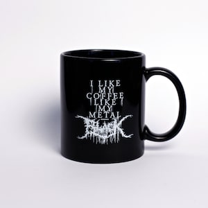 May include: Black ceramic coffee mug with a curved handle. The mug features white text that reads "I LIKE MY COFFEE LIKE MY METAL" with a stylized logo below. The mug is set against a white background.