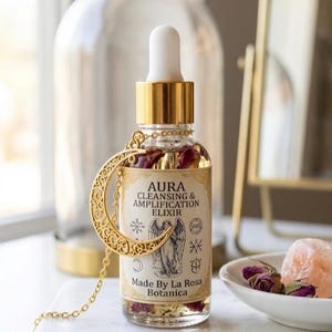May include: A clear glass dropper bottle filled with liquid and dried rosebuds, featuring a label that reads AURA CLEANSING & AMPLIFICATION ELIXIR Made By La Rosa Botanica. A gold-toned crescent moon pendant on a chain hangs around the bottle neck.
