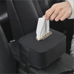 May include: A black, rectangular tissue box with a silver-colored trim around the tissue opening. White tissues are pulled from the box. The box is placed on a car's center console.