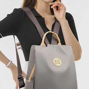 May include: A gray backpack with a gold handle and a small gold emblem. The backpack has brown straps and a side pocket. The bag is made of a smooth material and has a simple, modern design.