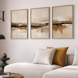 Puede incluir: Un conjunto de tres láminas de arte mural verticales enmarcadas con composiciones de paisajes abstractos en tonos beige, blanco, naranja quemado y negro. La obra muestra pinceladas horizontales superpuestas y texturas.