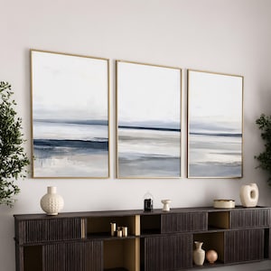 May include: A triptych of three vertical rectangular wall art pieces featuring an abstract landscape in shades of white, grey, and navy blue, each enclosed in a thin gold frame, displayed above a dark brown wooden sideboard with slatted doors.