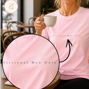 Könnte beinhalten: Hellrosa T-Shirt mit dem Text "Fictional Men Only." Das Shirt besteht aus einem weichen Material. Eine Person hält eine beige Tasse. Der Hintergrund zeigt ein Café mit Holzstühlen und -tischen.