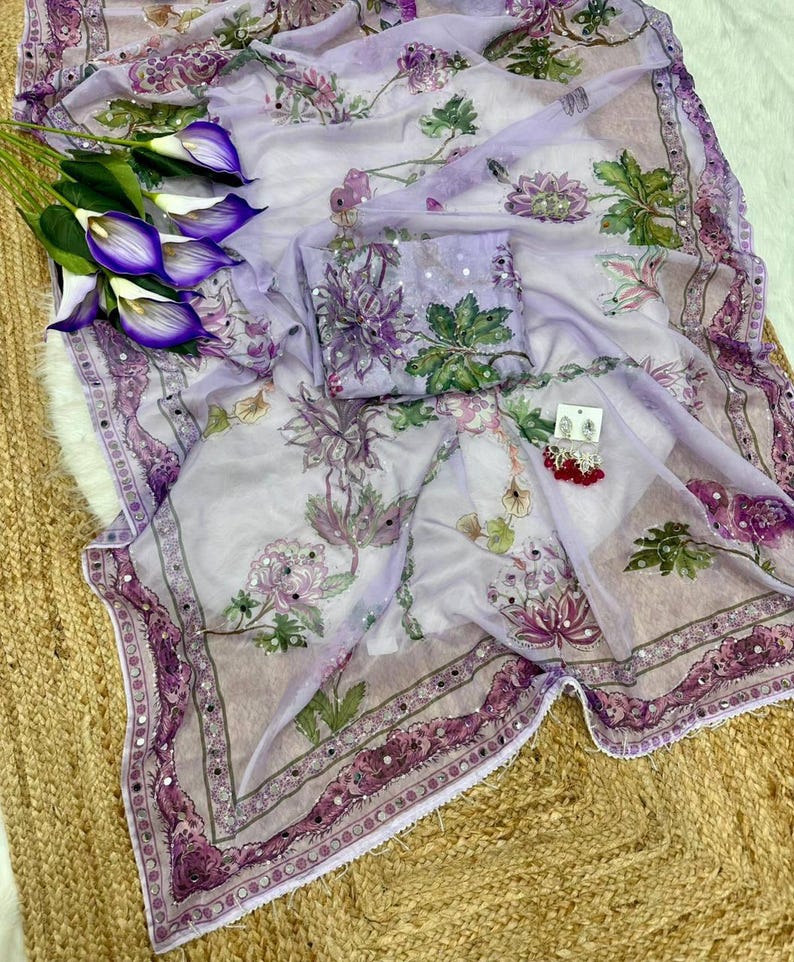 May include: A sheer, lavender-colored fabric with a floral pattern in shades of purple and green. The fabric is adorned with small, reflective embellishments and a decorative border. A matching pouch and earrings are also present.