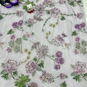 May include: A light purple fabric with embroidered floral patterns in shades of purple, pink, and green. The design includes large flowers, smaller blossoms, and leaves, with scattered sequins for added detail. The fabric is likely used for clothing or home decor.