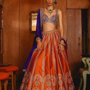 Op de afbeelding: Een vrouw in een traditionele Indiase outfit. De outfit bestaat uit een oranje rok met zilveren borduursel, een paarse en gouden top en een bijpassende paarse sjaal. De outfit is voorzien van sieraden.