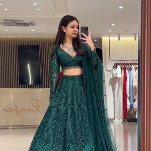 May include: A dark emerald green lehenga set. The set includes a long-sleeved, cropped top with a plunging neckline, a matching high-waisted skirt, and a sheer dupatta. The outfit is embellished with intricate patterns and sequins. A silver necklace is worn.