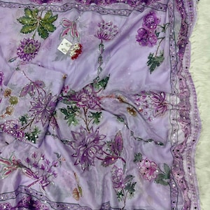 May include: A light purple floral scarf with a matching pouch. The scarf features a repeating pattern of purple flowers and green leaves. The edges are trimmed with a dark purple border and small silver embellishments. The fabric appears to be lightweight and flowing.
